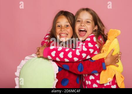 Les filles en pyjama coloré à pois contiennent de drôles oreillers lumineux. Les enfants aux visages heureux et aux cheveux amples. Enfants en vêtements brillants sur fond rose. Concept d'enfance, d'amitié et de bonheur Banque D'Images