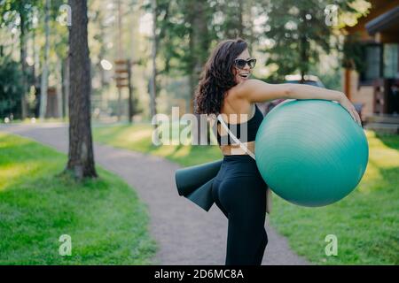 Photo extérieure de la femme heureuse brune porte le fitball et le tapis de fitness, porte des lunettes de soleil et des vêtements de sport, pose dans le parc vert, étant en bonne forme physique Banque D'Images