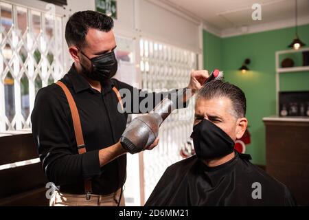 white caucasien barber peignes et souffle-drys cheveux d'un autre homme dans un salon de coiffure pendant la pandémie de coronavirus. ils vont rétro style hipster et nous tous les deux Banque D'Images