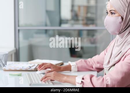 Jeune femme d'affaires contemporaine en hijab et masque de protection travaillant par bureau Banque D'Images