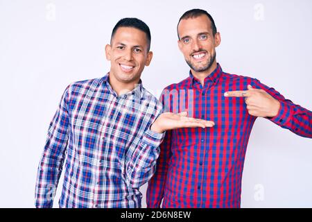 Jeune gay couple de deux hommes portant des vêtements décontractés stupéfaits et souriant à l'appareil photo tout en présentant avec la main et pointant avec le doigt. Banque D'Images