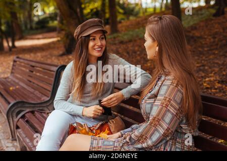 Portrait extérieur de deux jeunes femmes parlant assis sur un banc dans le parc d'automne. Des amis traînent ensemble. Banque D'Images