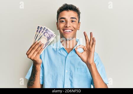 Jeune homme afro-américain élégant tenant 5000 billets de yens japonais signe OK avec les doigts, souriant sympathique gestante excellent symbole Banque D'Images