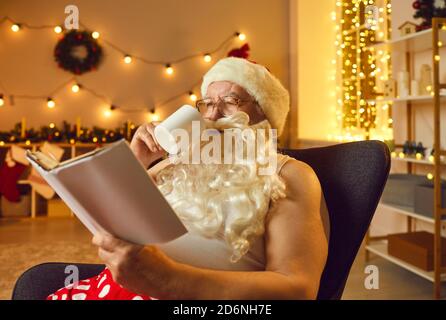 Le père Noël lit un livre et boit du café assis dans un fauteuil confortable. Banque D'Images