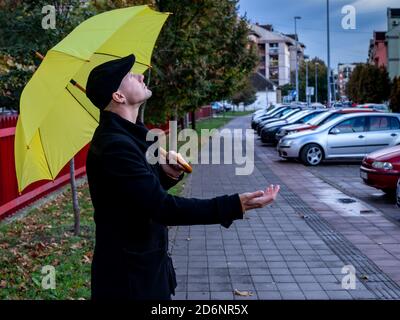 Homme en manteau noir, debout dans les rues de la ville sous le parapluie jaune vérifiant avec la paume de sa main si la pluie avait cessé de tomber. Prévisions. Banque D'Images