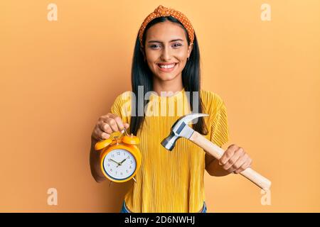 Jeune femme brune tenant un réveil et un marteau souriant avec un sourire heureux et frais sur le visage. Montrant les dents. Banque D'Images