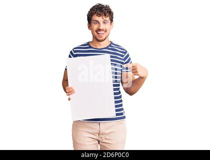 Jeune homme beau avec cheveux bouclés tenant une bannière vide sourire sourire sourire sourire heureux avec la main et le doigt Banque D'Images