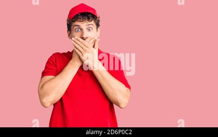 Jeune homme beau avec cheveux bouclés portant l'uniforme de livraison choqué couvrant la bouche avec les mains pour erreur. Concept secret. Banque D'Images