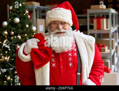 Le Père Noël heureux portant un costume tenant un sac sac avec des cadeaux dans l'atelier. Banque D'Images