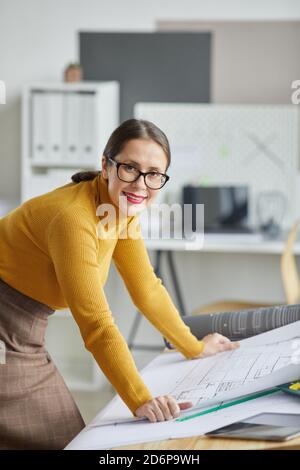 Portrait vertical d'une femme architecte adulte souriant à l'appareil photo s'appuyer sur un bureau de dessin au bureau et travailler sur des plans Banque D'Images