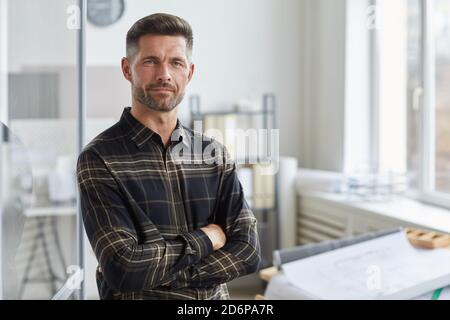 Portrait à la taille de l'architecte barbu confiant regardant l'appareil photo tout en se tenant debout avec les bras croisés par le bureau de dessin, dans l'espace de copie Banque D'Images