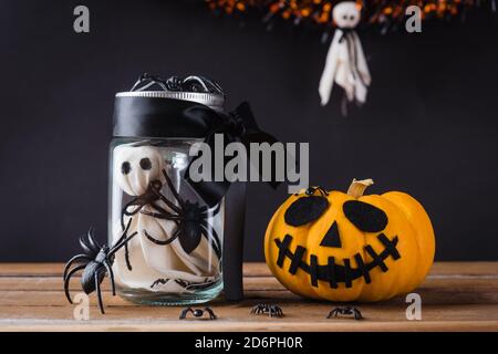 Drôle Halloween jour décoration fête, le fantôme blanc visage effrayant et araignée noire en verre pot et potiron dehors côté sur table en bois, studio photo isola Banque D'Images