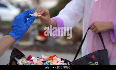 Halloween 2020 : gros plan d'une main gantée donnant des bonbons à une jeune fille dans un costume Banque D'Images