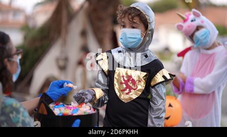 Les enfants portant des costumes et des masques de visage hors de trick ou de traiter sur Halloween 2020. Une personne remet des bonbons avec une main gantée. Banque D'Images