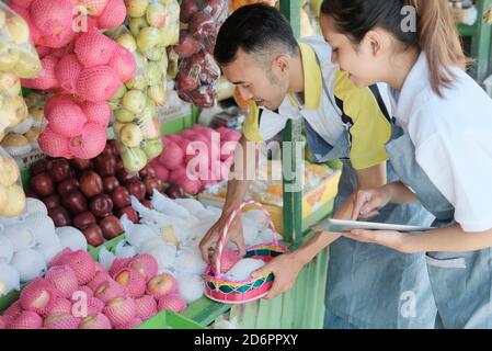 vendeur femelle utilisant une tablette et vendeur mâle se tient près d'un affichez les fruits frais tout en préparant les fruits sur le panier à faites un paquet de colis Banque D'Images