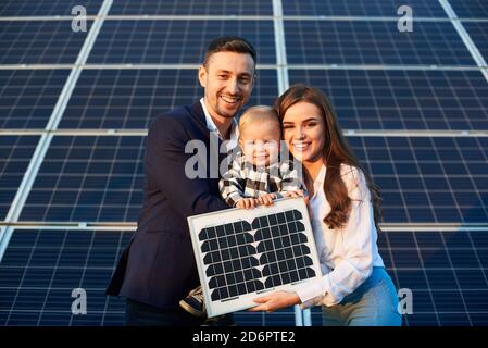 Portrait d'une jeune famille tenant un petit panneau solaire et un bébé garçon, souriant et regardant l'appareil photo. Concept de génération verte Banque D'Images