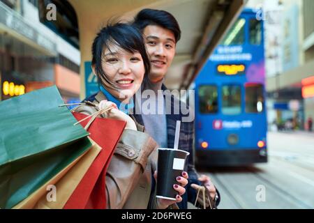 un jeune couple asiatique heureux attend le bus dans une ville moderne avec des sacs à provisions à portée de main Banque D'Images