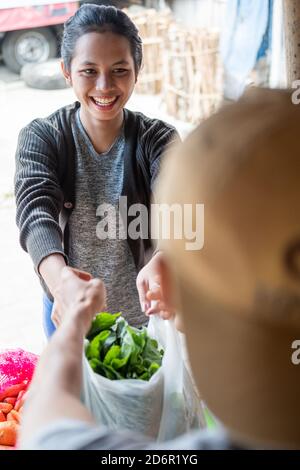 Gros plan d'une femme asiatique souriante tenant un sac en plastique rempli de légumes tout en faisant des achats dans un marché traditionnel Banque D'Images