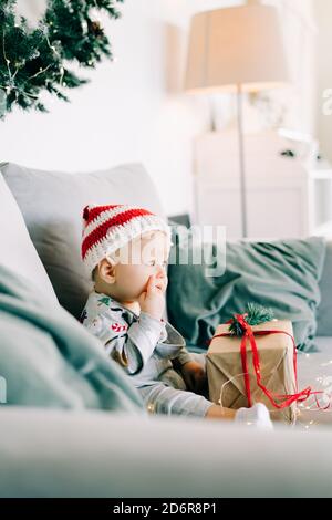 Le petit garçon couvant est assis dans un chapeau de père Noël et un costume de vacances sur un canapé gris avec un grand cadeau avec ruban rouge, contre Mur blanc avec couronne de sapin naturel.CH Banque D'Images