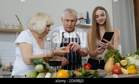 Les grands-parents couplent des légumes pour les salades dans la cuisine. Homme mature et femme écoutant la recette, conseils de petite fille avec tablette numérique. Alimentation saine. Régime alimentaire cru Banque D'Images