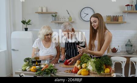 Les grands-parents couplent des légumes pour les salades dans la cuisine. Homme mature et femme écoutant la recette, conseils de petite fille avec tablette numérique. Alimentation saine. Régime alimentaire cru Banque D'Images