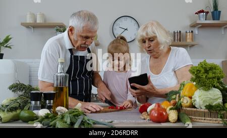 Les grands-parents âgés s'accouplent avec une petite-fille qui coupe des légumes pour la salade dans la cuisine. Homme et femme matures écoutant la recette, conseils de la tablette numérique. Alimentation saine. Régime alimentaire cru Banque D'Images