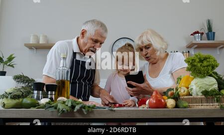 Les grands-parents se couple avec la petite-fille qui coupe des légumes pour les salades dans la cuisine. Homme et femme matures écoutant la recette, conseils de la tablette numérique. Alimentation saine. Régime alimentaire cru Banque D'Images