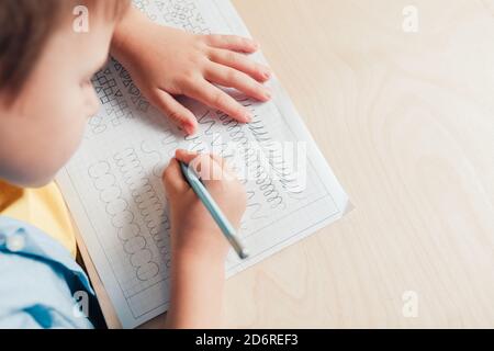 Gros plan sur un adorable garçon qui fait ses devoirs. Écriture enfant avec crayon. Pratique de préécriture pour préparer les mains pour écrire des lettres. Concept d'éducation des enfants Banque D'Images