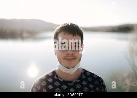 Jeune touriste dans le masque facial et les lunettes debout près de la rivière derrière les montagnes tout en regardant l'appareil photo dans l'arrière éclairé Banque D'Images