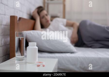 Pot de pilules et verre d'eau sur table de chevet et jeune femme dépressive couché sur le lit en arrière-plan, espace de copie Banque D'Images