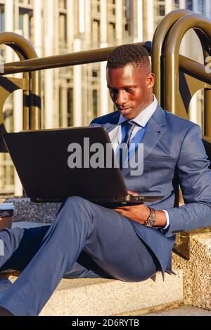 Vue du corps entier du gestionnaire financier afro-américain concentré dans élégant bleu costume formel assis sur les escaliers près de moderne bâtiment et lecture Banque D'Images