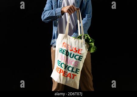 Rognez l'acheteur mâle anonyme tenant le sac de shopping en tissu avec le texte message de réutilisation et de recyclage sur fond noir Banque D'Images
