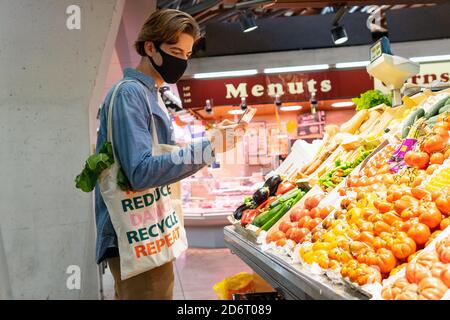 Jeune homme client dans un masque de protection transportant écologique recyclé sac à provisions en tissu et choix de produits frais tout en prenant des photos sur smartph Banque D'Images