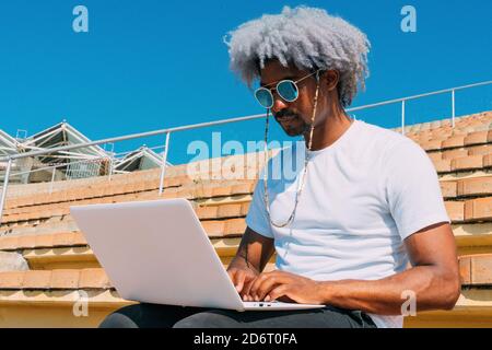 Afro et homme noir avec des lunettes de soleil travaillant avec son ordinateur portable dans la rue. Travailler avec l'ordinateur portable à l'extérieur. Homme noir travaillant sur l'ordinateur. Banque D'Images