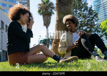 Des jeunes Afro-américains millénaires positifs dans des tenues tendance qui prennent en compte assis sur l'herbe dans le parc Banque D'Images