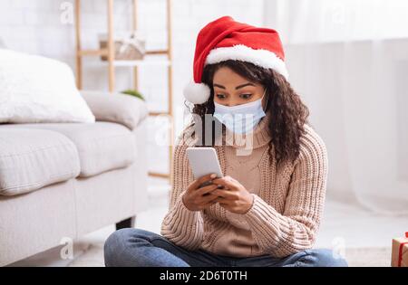 Bonne femme noire en chapeau de père noël et masque en utilisant le téléphone cellulaire Banque D'Images