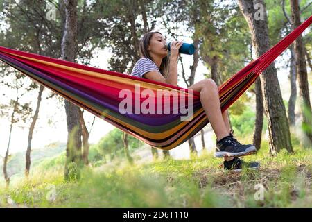 Vue latérale à angle bas d'une femme assoiffée en suspension hamac dans le parc et profiter de l'eau douce à la bouteille jour d'été Banque D'Images
