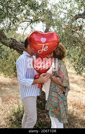 Vue latérale d'un couple méconnaissable debout près de l'arbre jardiner et se cacher derrière des ballons d'air rouges en forme de cœur Banque D'Images