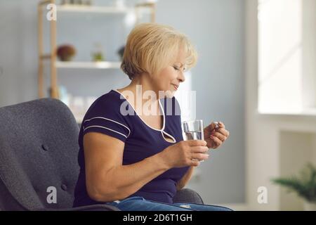 Femme mûre souriante assise dans un fauteuil et prenant une pilule médicale et de l'eau pure dans le verre dans les mains Banque D'Images