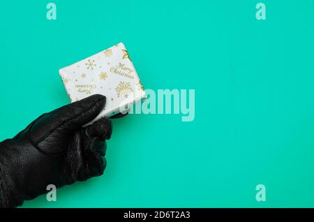 Main dans le cuir noir gant contient le cadeau de Noël enveloppé sur fond turquoise. Petite boîte rectangulaire enveloppée de papier d'emballage blanc. Noël Banque D'Images