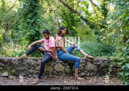 Vue latérale d'une jeune femme et d'un homme assis dos à dos dans un parc vert et en parcourant les smartphones Banque D'Images
