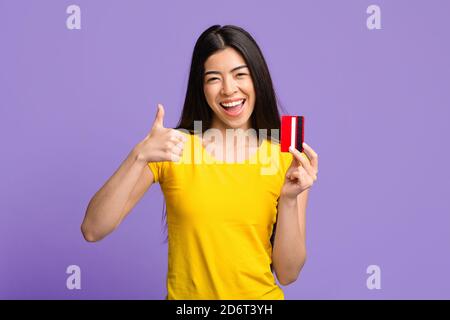 Une femme asiatique joyeuse et enthousiaste tenant une carte de crédit et montrant Thumb Haut Banque D'Images