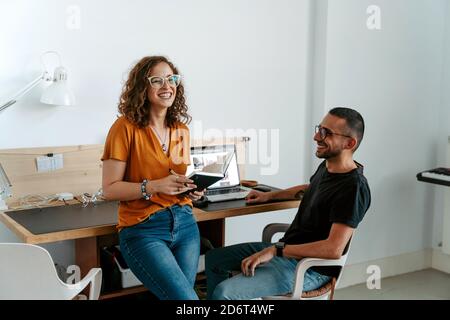 Jeune femme concentrée avec des cheveux bouclés dans des vêtements décontractés et des lunettes qui prennent des notes tout en travaillant avec un collègue dans moderne Banque D'Images
