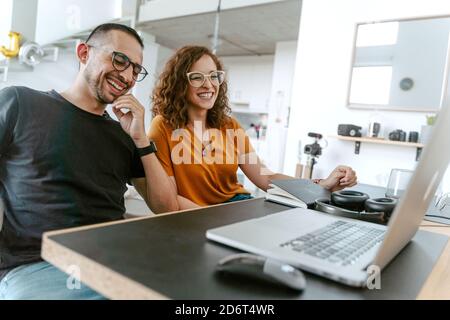Joyeux jeune couple divers en vêtements décontractés et lunettes de vue pendant assis ensemble et passer des appels vidéo sur un ordinateur portable Banque D'Images
