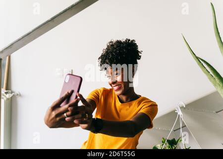 Femme millénaire afro-américaine excitée avec une tenue décontractée et un selfie avec téléphone mobile et rire tout en se reposant dans un appartement moderne Banque D'Images