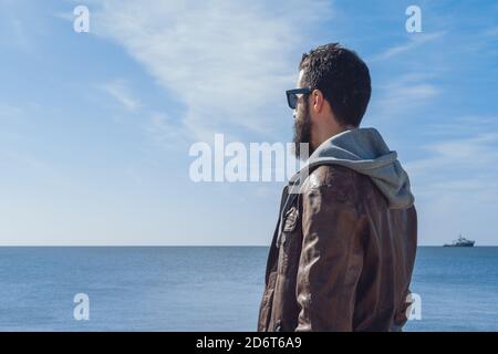 Vue latérale du voyageur masculin pensif à barbe ethnique dans la mode vêtements et lunettes de soleil se détendant sur la plage rocheuse de la mer dedans Sept Sœurs Banque D'Images