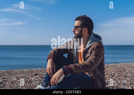 Vue latérale du voyageur masculin pensif à barbe ethnique dans la mode vêtements et lunettes de soleil se détendant sur la plage rocheuse de la mer dedans Sept Sœurs Banque D'Images