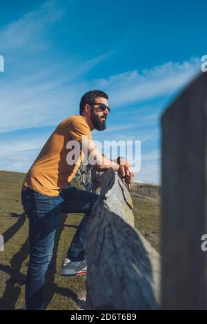 Vue latérale d'un jeune homme ethnique décontracté avec une tenue décontractée et des lunettes de soleil se penchent sur une clôture en bois et admirent la nature contre ciel bleu Banque D'Images