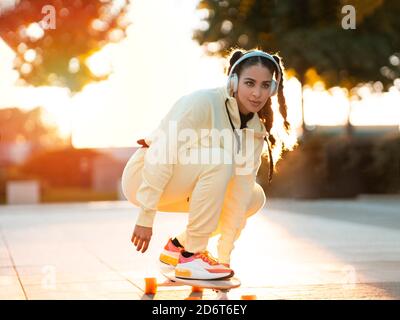 Tout le corps de jeune femme ethnique positive dans des vêtements de sport élégants monter à bord de la longboard et écouter de la musique dans les écouteurs du parc au coucher du soleil Banque D'Images