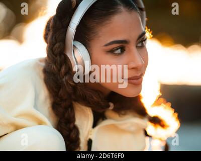 Courte positive jeune femme ethnique dans l'élégant sportswear de cheval long-board et écouter de la musique dans un casque au parc au coucher du soleil Banque D'Images